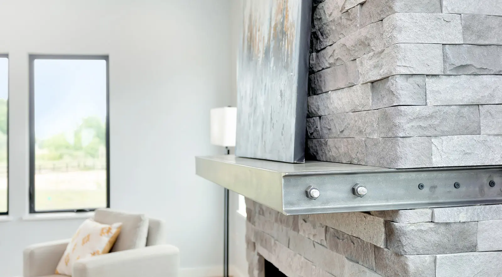 Close-up of a modern stone fireplace with a metal mantel and a large painting on top; this bright, minimalist living room showcases Walla Walla custom home design with a beige armchair and two windows.