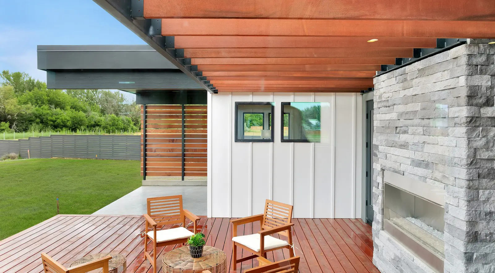 Modern patio with wooden deck, chairs, and table, a stone fireplace, and a pergola roof. Featuring Walla Walla custom home design touches, the space boasts a white paneled wall with windows and a green lawn in the background.