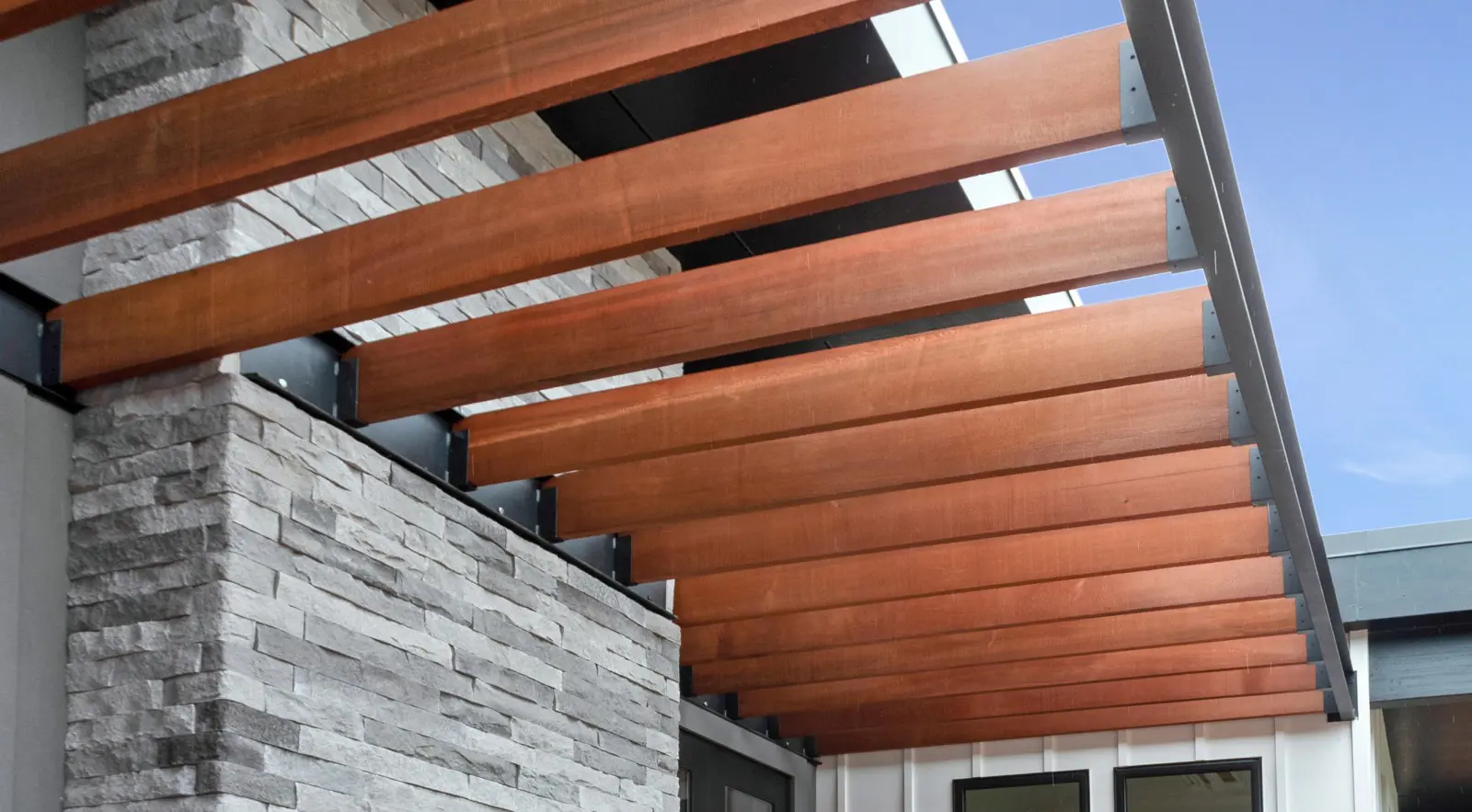 Wooden pergola beams extend from a modern building with stone veneer siding, a hallmark of Walla Walla custom home design, creating partial shade above windows under a clear blue sky.