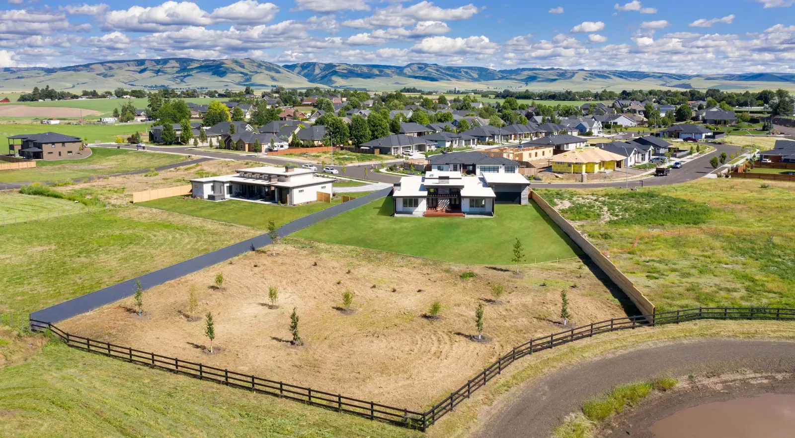 Aerial view of a modern Walla Walla custom home design with a large yard and fenced property in a suburban neighborhood, surrounded by fields and distant hills under a partly cloudy sky.