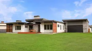 Modern single-story house with flat roofs, large windows, and stone and wood accents—a stunning example of Walla Walla custom home design—set on a spacious green lawn under a blue sky.