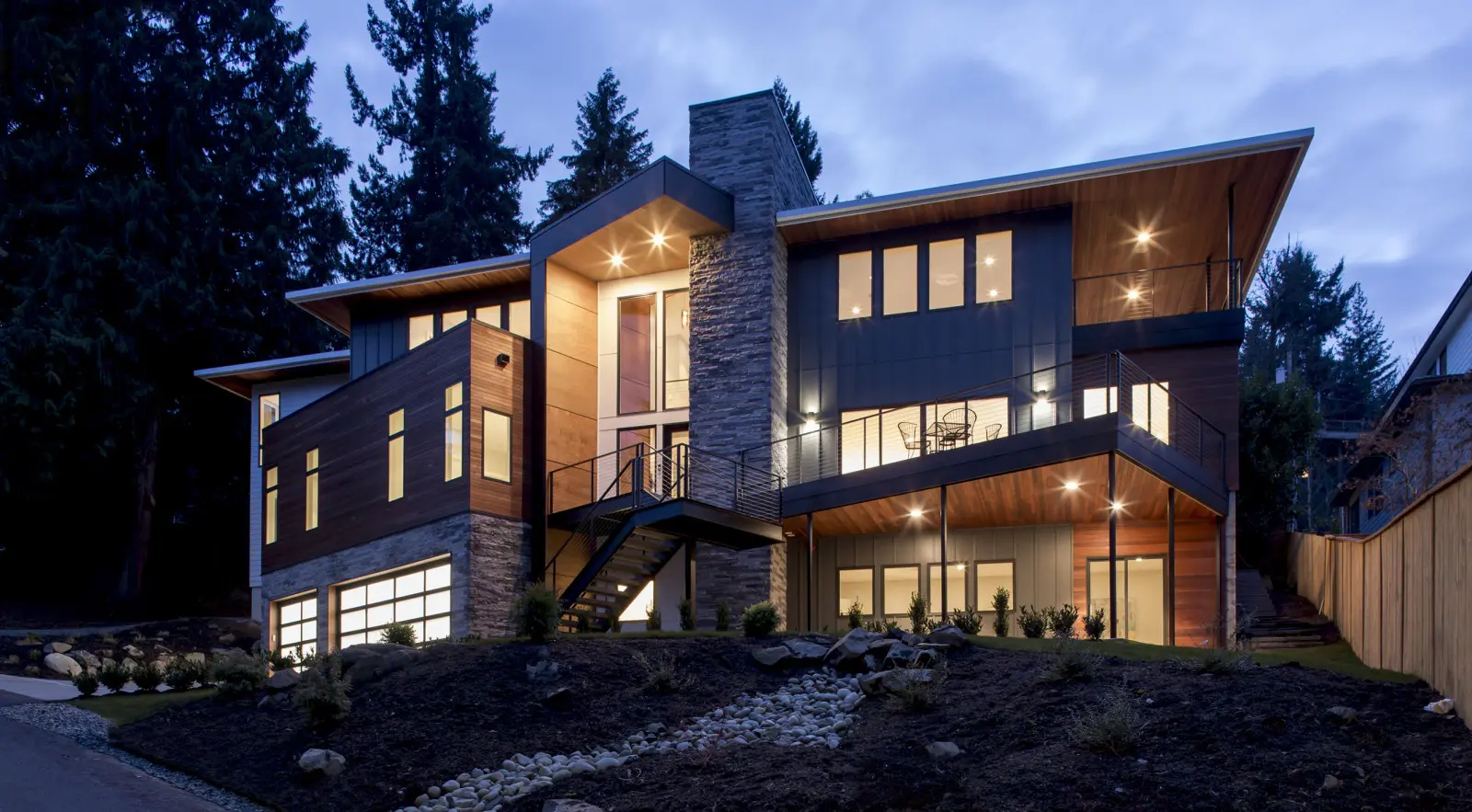 Modern two-story Bellevue Meydenbauer custom home design featuring large windows, stone and wood exterior, outdoor lighting, and a sloped yard surrounded by trees at dusk.