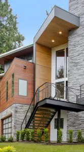 Modern Bellevue Meydenbauer custom home design features a two-story exterior with mixed wood and stone siding, large windows, metal stair railing, and a beautifully landscaped front yard.