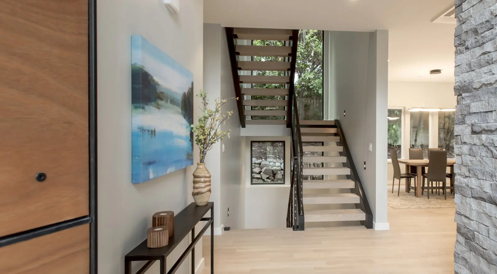 Modern home entryway with light wood floors, a console table with decor, and open-riser staircase—showcasing Bellevue Meydenbauer custom home design—with a view into the dining area featuring large windows.
