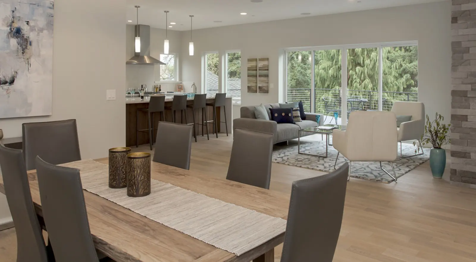 Modern open-concept living space featuring Bellevue Meydenbauer custom home design, with a dining table in the foreground, a kitchen with barstools, and a seating area near large windows overlooking lush greenery.