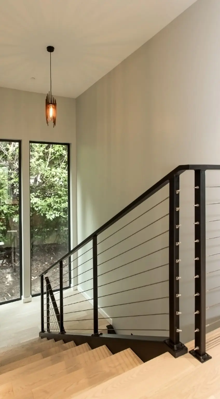A modern staircase with light wood steps and a black cable railing enhances this Bellevue Meydenbauer custom home design, set next to large windows overlooking greenery beneath a contemporary pendant light.