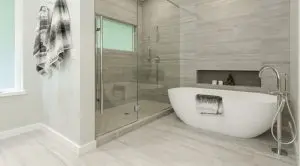Modern bathroom in a Bellevue Meydenbauer custom home design, featuring a glass-enclosed shower, freestanding white bathtub, wall-mounted fixtures, and gray towels hanging and folded.