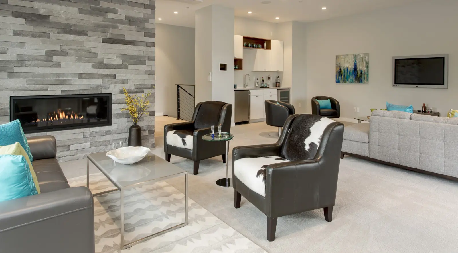 Modern living room with a stone fireplace, black armchairs, gray sofa, glass coffee table, wall-mounted TV, and a small bar area—showcasing Bellevue Meydenbauer custom home design.
