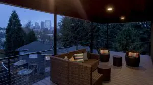 Covered outdoor patio with wicker furniture and round side tables, offering a city skyline view through trees at dusk—showcasing Bellevue Meydenbauer custom home design.