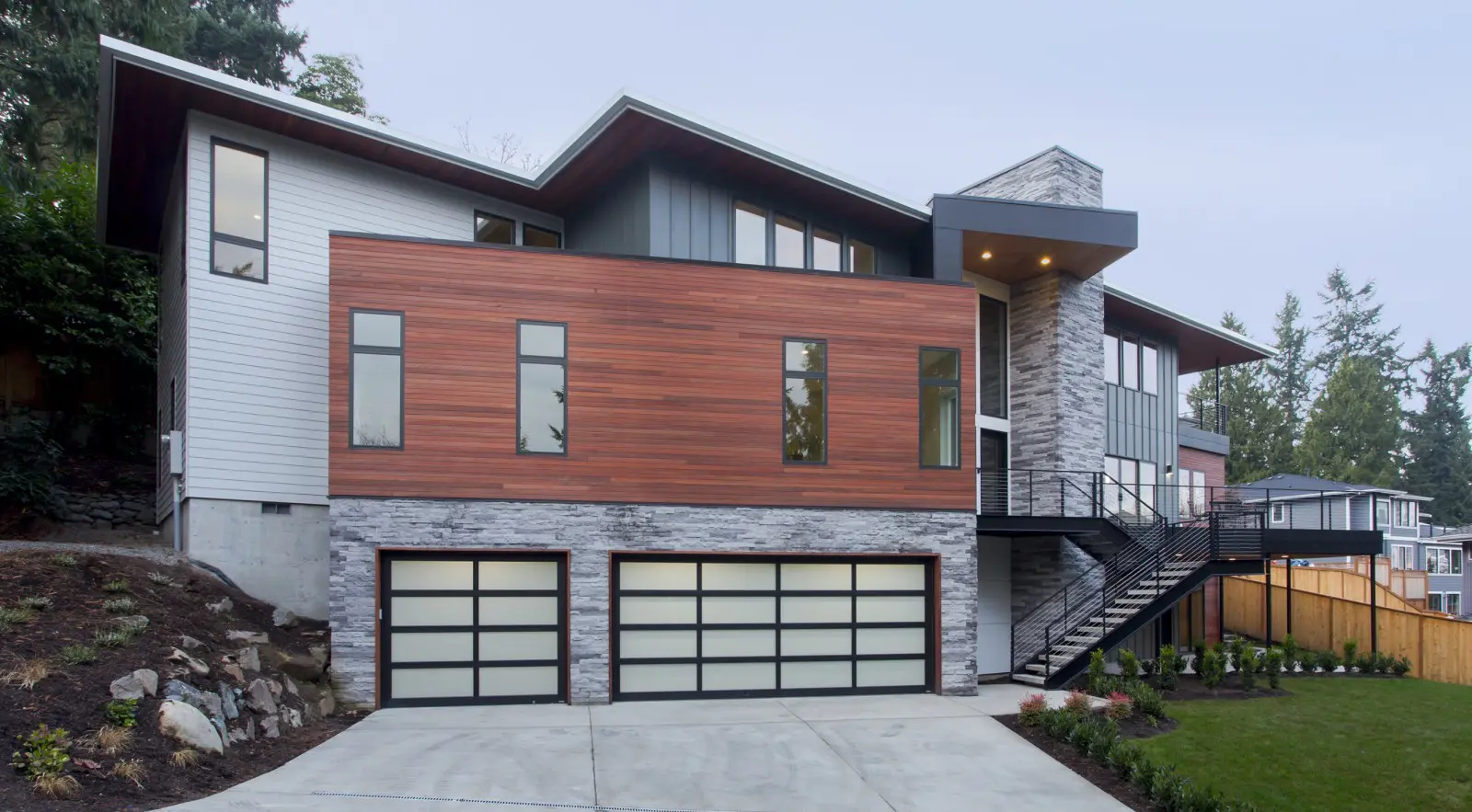 Modern two-story Bellevue Meydenbauer custom home design features a mix of wood, stone, and metal exterior, large windows, a three-car garage, and a striking black metal staircase.