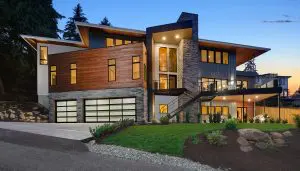 Modern two-story house with large windows, wood and stone exterior, three-car garage, and landscaped front yard at dusk, exemplifying Pacific Northwest luxury residential architecture.