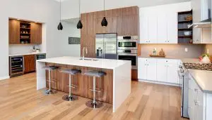 Modern kitchen with wood floors, an island with three stools, stainless steel appliances, white and wood cabinets, and pendant lights—showcasing elements of Pacific Northwest luxury residential architecture.