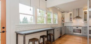 Modern kitchen remodels feature light gray cabinets, stainless steel appliances, a long counter with bar stools, and large windows providing natural light.