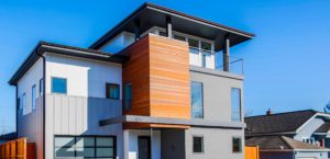 Modern two-story new construction house with a mix of white, gray, and wood paneling, large windows, and a rooftop balcony, set against a clear blue sky.