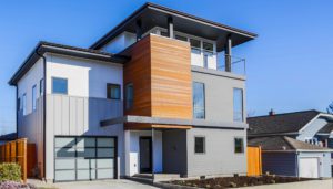 Modern two-story house featuring West Seattle custom home design, with gray siding, wood accent panel, large windows, a flat roof, and an attached garage on a clear day.
