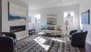 Modern living room in a West Seattle custom home design featuring a patterned rug, two armchairs, a sofa, glass coffee table, wall art, and built-in fireplace. Natural light enters through three windows.