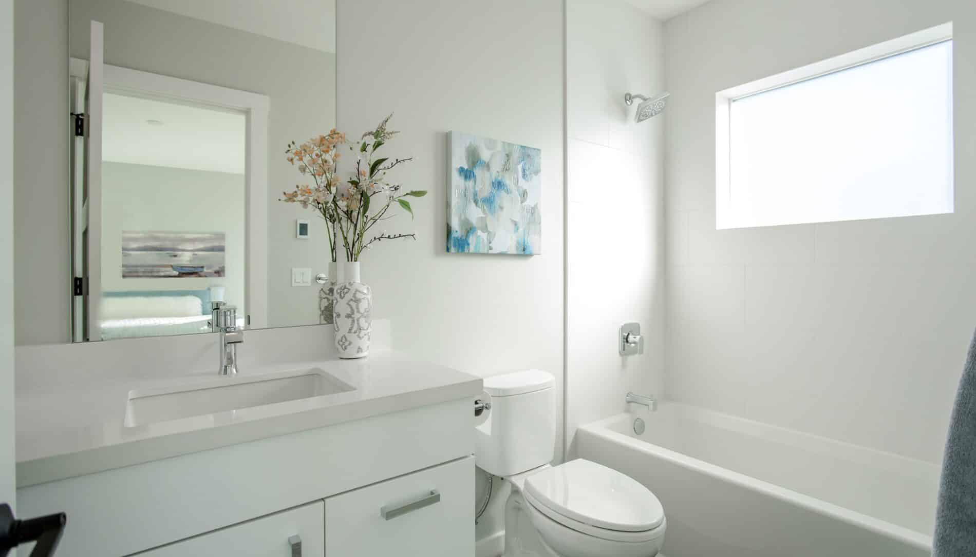 Modern white bathroom in a West Seattle custom home design, featuring a sleek vanity, sink, toilet, bathtub, stylish wall art, vase with flowers, and a frosted window that fills the space with natural light.