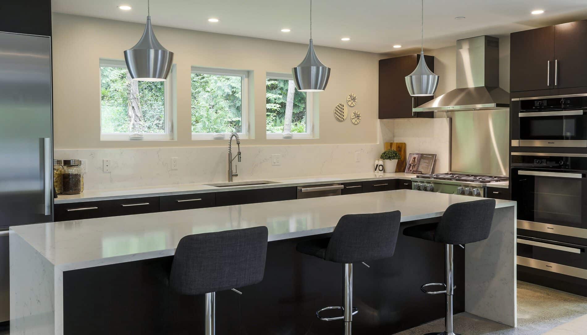 Modern kitchen featuring dark cabinets, stainless steel appliances, a large island with three black barstools, pendant lights, and three windows above the sink—showcasing elegant Mercer Island custom home design.