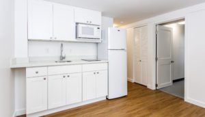 A small, modern kitchen with white cabinets, a countertop sink, a fridge, a microwave, and a stove, set on a wood floor next to a white door and a closet with louvered doors.