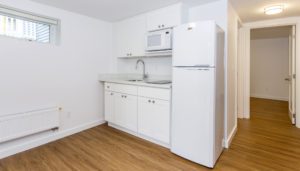 Small kitchen with white cabinets, a sink, microwave, and refrigerator. The floor is wood, and there is a window and a hallway visible.