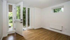 Empty room with white walls, wood-style flooring, a small window, radiator, and a glass door open to a garden area.