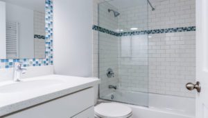 Modern bathroom with a white vanity, a sink, a toilet, and a bathtub with a glass door. The shower has white subway tiles and a blue mosaic accent strip.