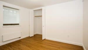 Empty room with white walls, wooden floor, a window with blinds, an open closet with a single shelf and rod, and a wall-mounted radiator.