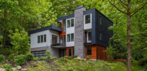 Modern new construction two-story house with stone, wood, and black panel exterior, surrounded by dense green trees and vegetation.