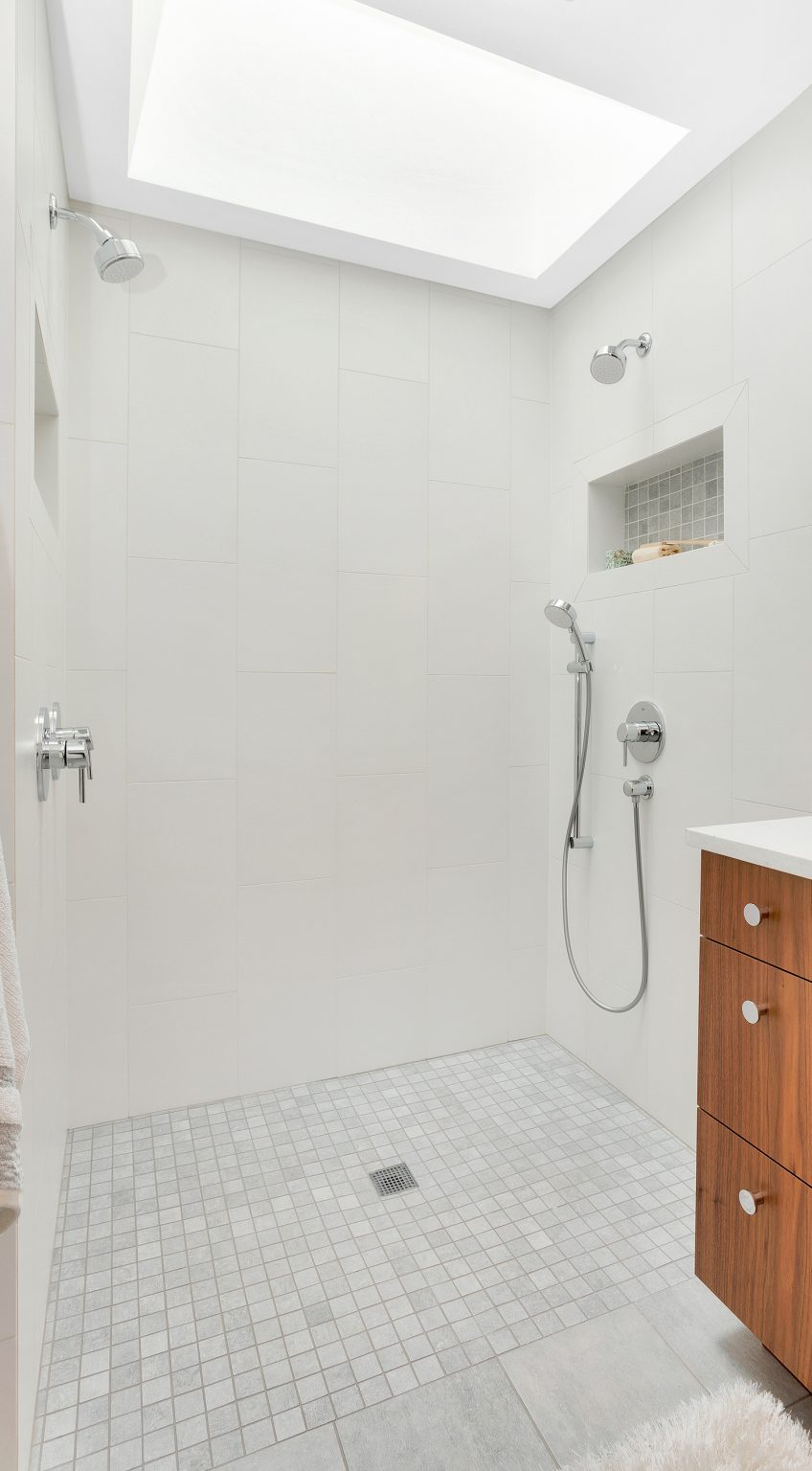 Modern walk-in shower with dual showerheads, built-in wall niches, light gray tiles, and a wooden vanity on the right—showcasing sleek Walla Walla custom home design.