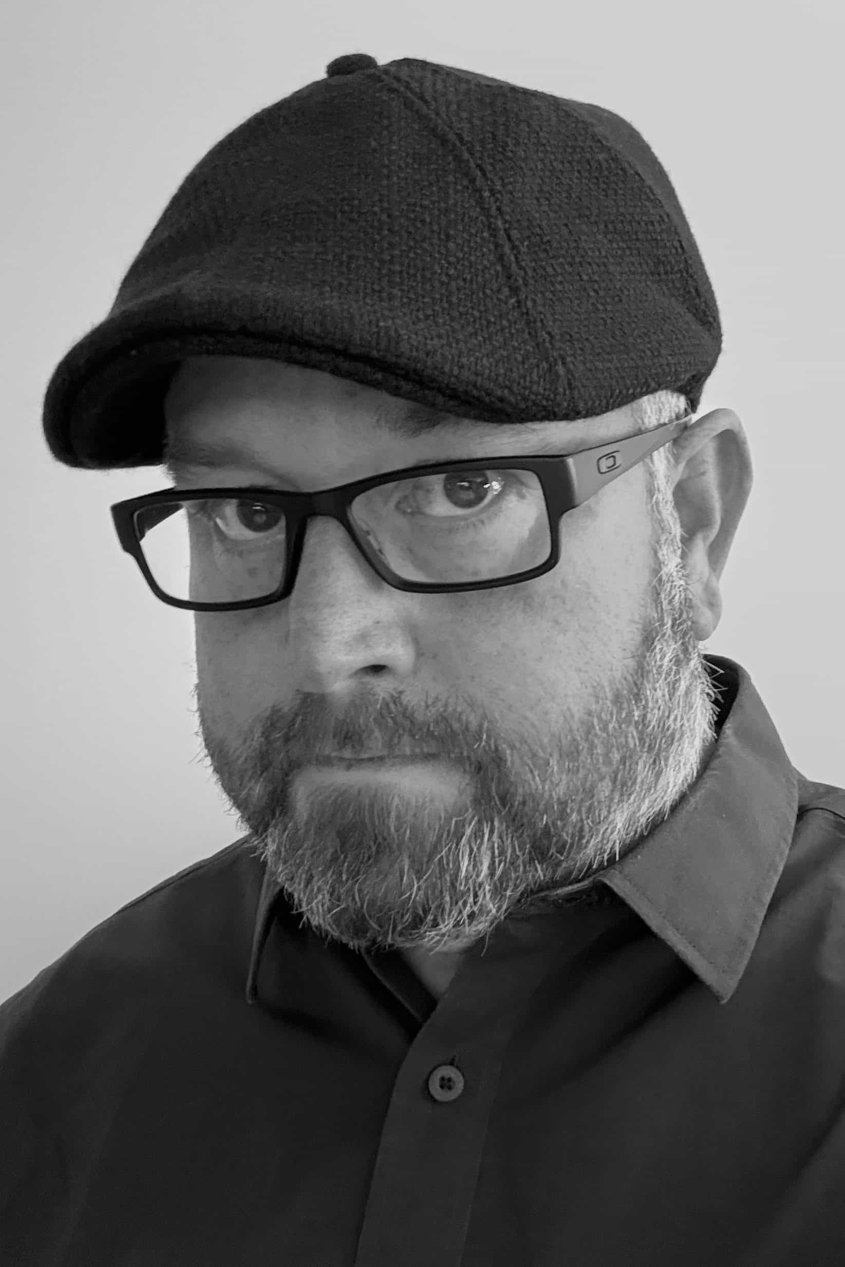 A bearded man wearing glasses, a flat cap, and a collared shirt—captured in black and white—looks toward the camera, evoking the thoughtful presence of a Seattle architect.